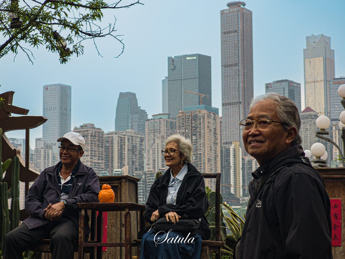 几位长者在河岸边并坐，身后是城市天际线。